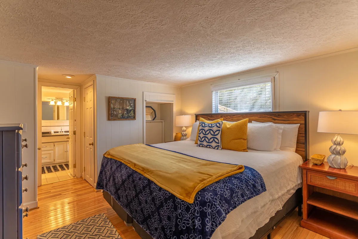 A serene bedroom with a plush bed adorned with decorative pillows, a nightstand with a glowing lamp, and tasteful artwork on the wall. The soft lighting and comfortable layout evoke a sense of tranquility, suggesting restful mornings and peaceful nights.