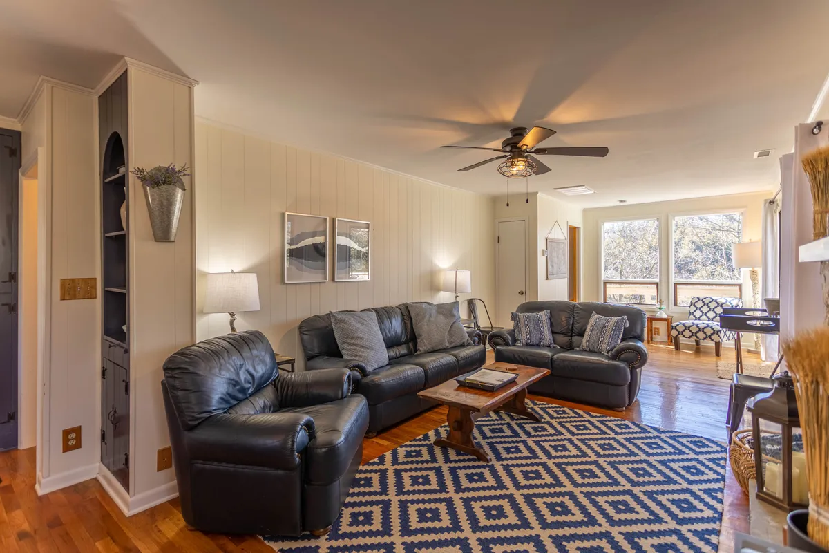 The image displays a welcoming living space with black leather sofas, a wooden coffee table perfect for games or snacks, a geometric patterned rug, and large windows that let in natural light. The arrangement invites gatherings and lively activities, emphasizing flexibility and comfort.