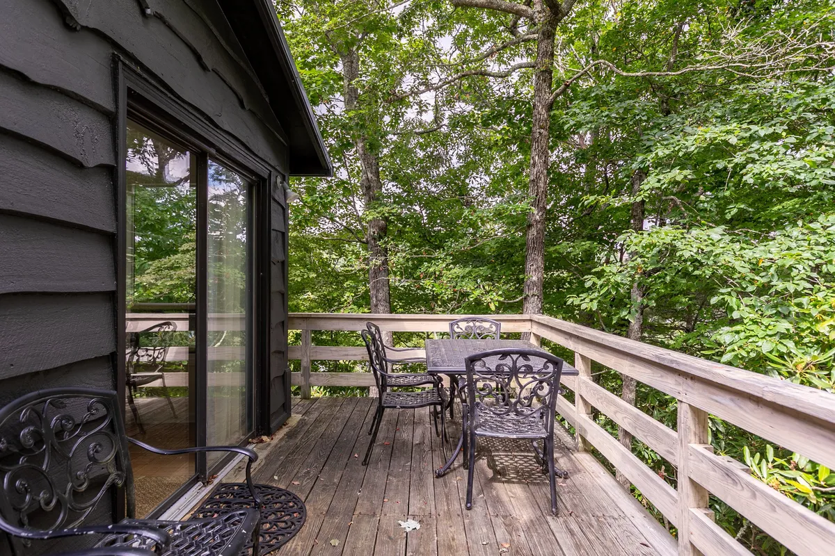 An inviting outdoor dining table with metal chairs sits on a spacious deck surrounded by greenery and a wooden railing. The view from the table is open to the lush landscape, offering a tranquil setting for meals or relaxation. The overall scene is peaceful and well-suited for gatherings or quiet moments.
