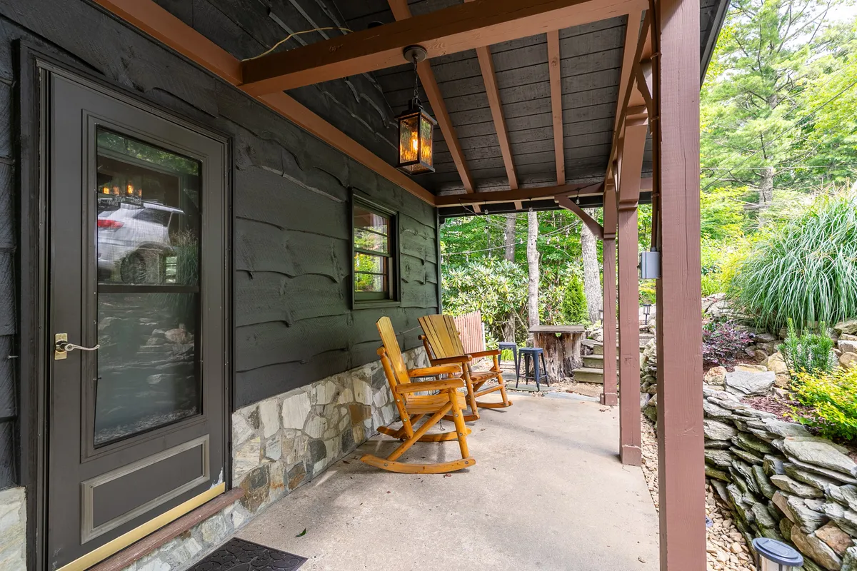 A cozy exterior porch features two rocking chairs, a stone wall, wooden beams, and hanging lanterns. Lush greenery surrounds the space, inviting guests to sit, relax, and enjoy the natural mountain setting. The mood is welcoming and tranquil, perfect for quiet moments or sharing with a pet.