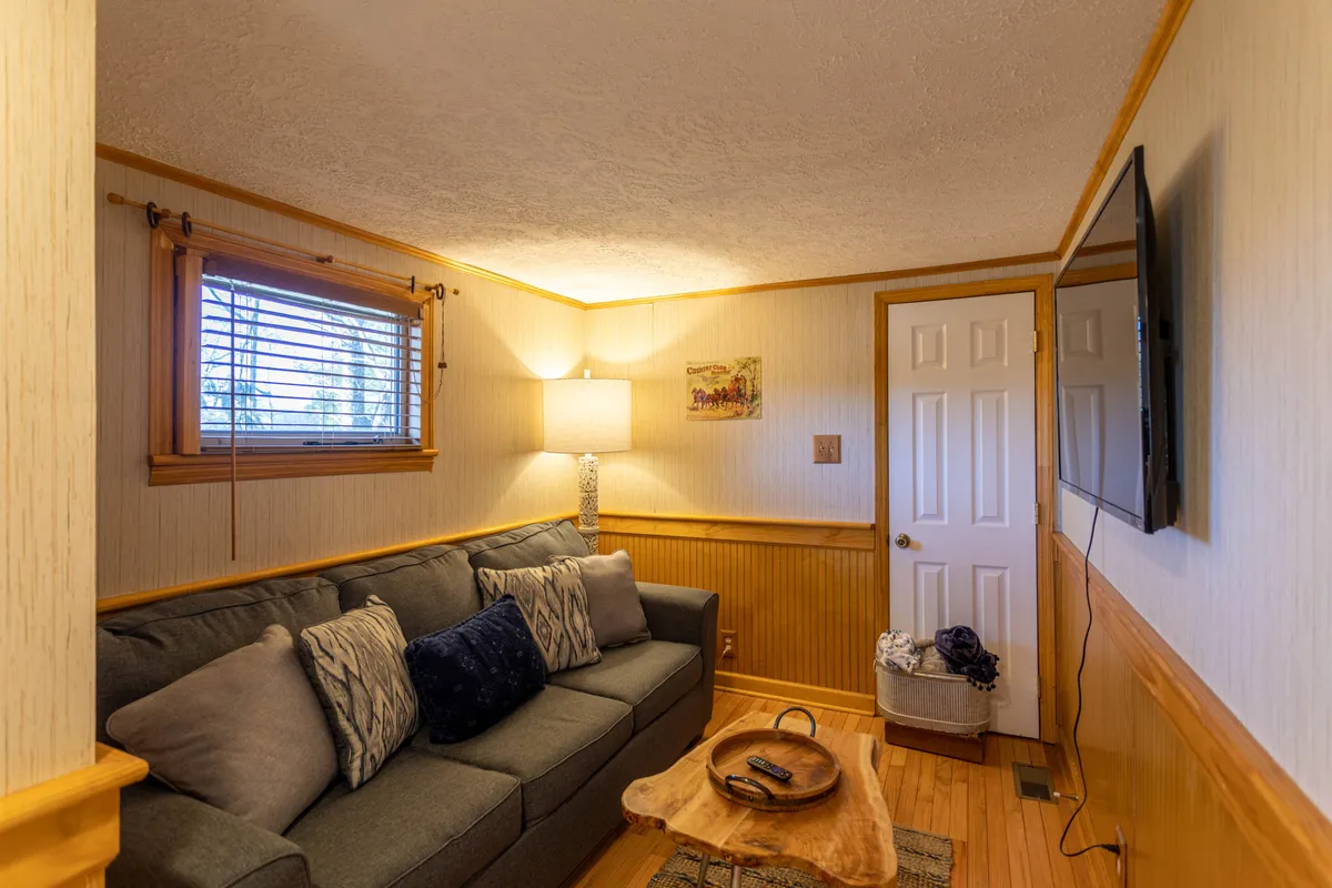This image showcases a cozy den area featuring a comfortable sofa with decorative cushions, a wooden coffee table, a lamp for ambient lighting, wall art, and a television positioned perfectly for movie nights or gaming sessions. The layout encourages both relaxation and socializing, making it ideal for group entertainment or quiet downtime.