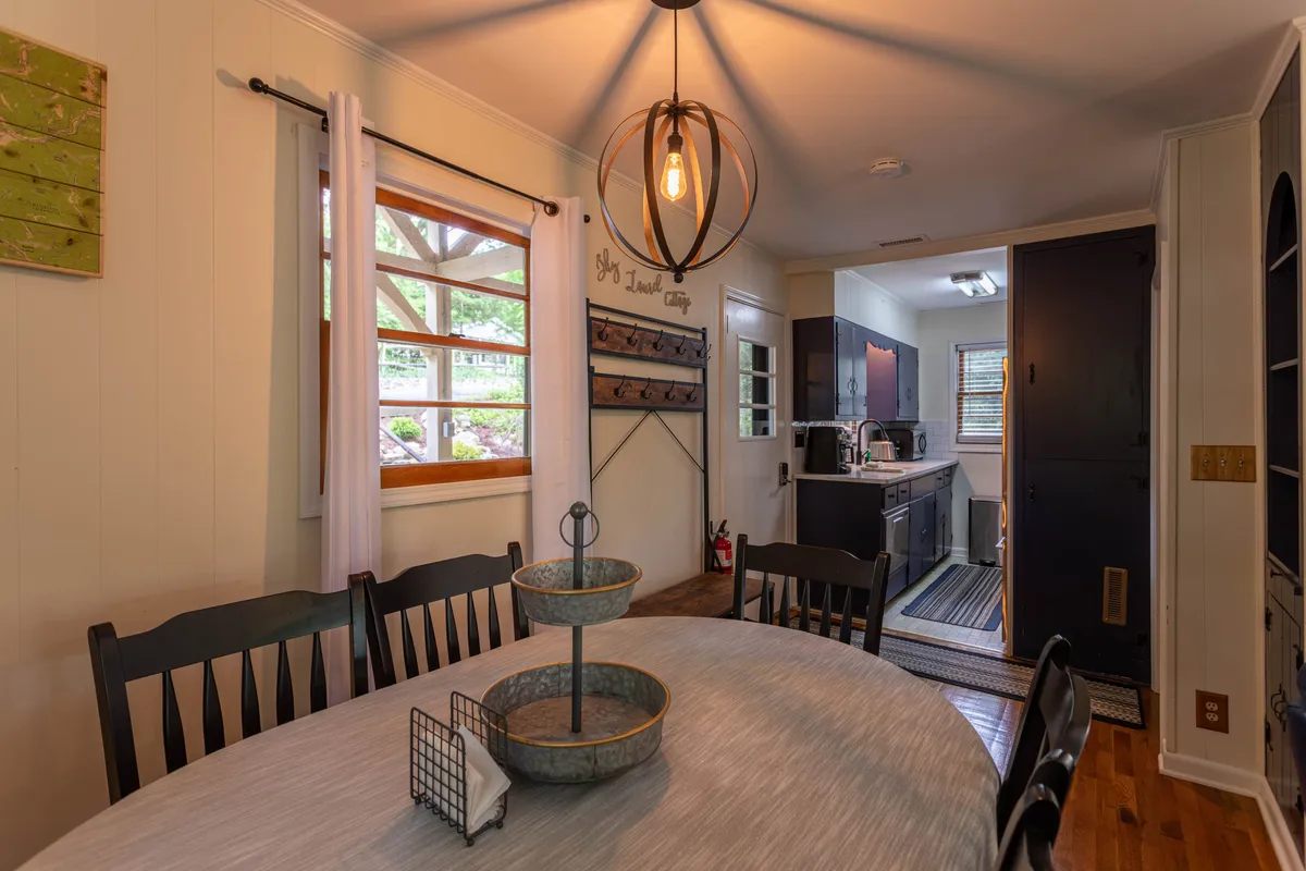 A cozy dining table situated under a stylish light fixture with curtains framing a nearby window. The table is adjacent to the kitchen entrance and decorated with tasteful wall art, suggesting a welcoming space for meals and socializing.
