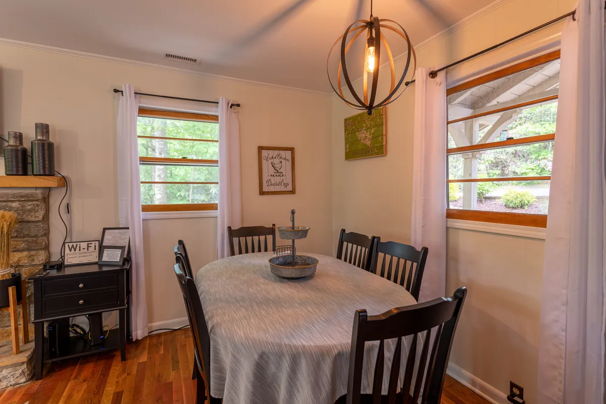 A dining area featuring an elegant table, a modern chandelier, large windows providing natural light, and a view of the cozy fireplace. Artistic wall decor and a spacious setting suggest a perfect spot for intimate meals or lingering conversations.