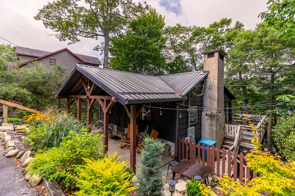 The image shows the wooden porch of Sky Laurel Cottage, complete with a prominent chimney and surrounded by a landscaped garden. The porch provides an inviting spot for relaxation, with views of the garden and the surrounding natural setting.