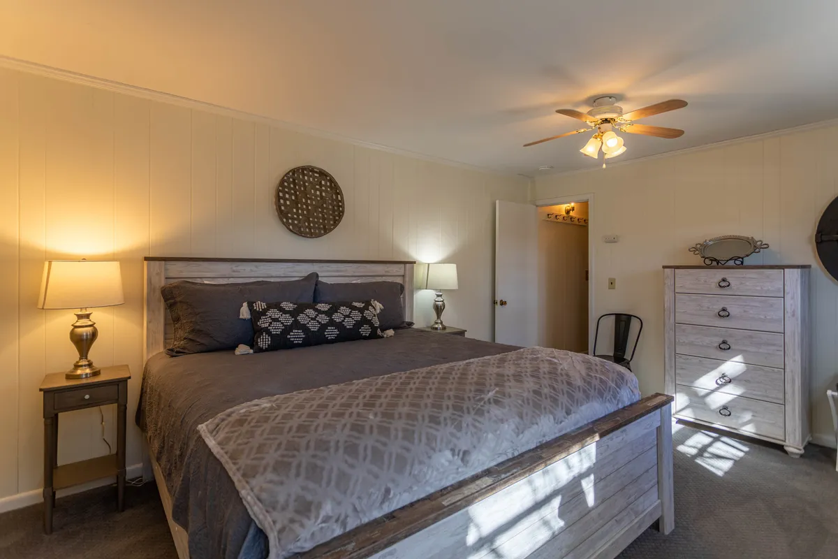 A tranquil bedroom with a bed topped with decorative pillows, a nightstand featuring a lamp, and a dresser. The room is filled with soft natural light, evoking a sense of calm and comfort—ideal for restful nights.