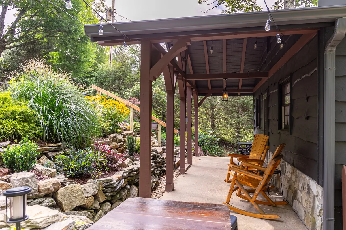 A cozy outdoor scene with wooden rocking chairs arranged on a landscaped garden patio, strung with warm hanging lights. The stone pathway and surrounding greenery create a welcoming space for outdoor gathering and relaxation.
