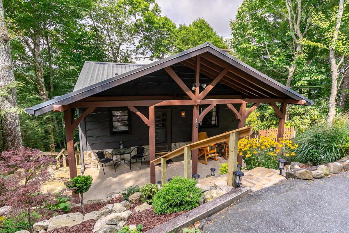 This exterior image showcases the inviting front porch of Sky Laurel Cottage, highlighting wooden beams, a rock garden, flower beds, and cozy seating. The scene is bathed in soft natural light, suggesting an ideal spot to unwind after exploring local trails with your pet.
