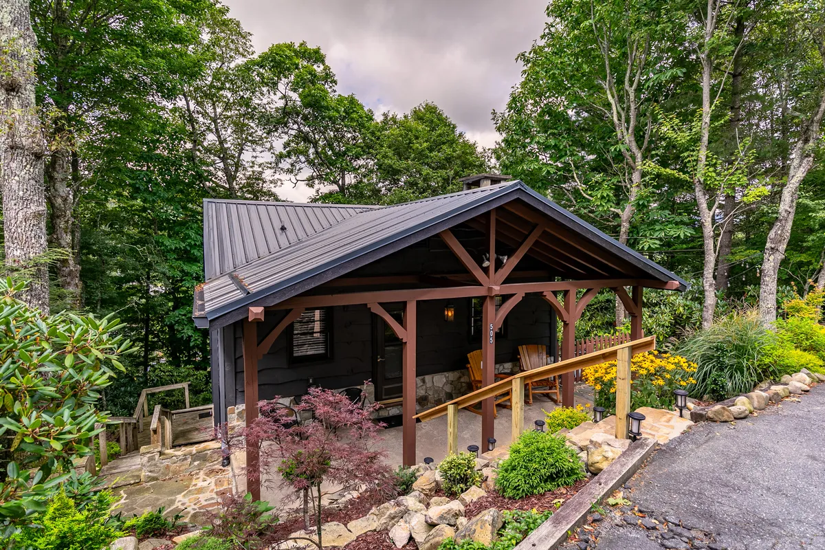 The image showcases the charming exterior of Sky Laurel Cottage, with a cozy front porch perfect for relaxing, a beautifully landscaped garden, and a stone pathway inviting guests to explore the outdoors. The natural setting is peaceful, surrounded by greenery and hinting at sunset views from the porch.